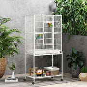 White bird cage with two birds beside large plants and supplies on the bottom shelf