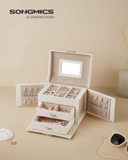 White jewellery box open on a beige dressing table beside sunglasses, filled with rings and beads