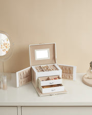 Open white jewellery box on a dressing table beside a round mirror and a perfume bottle, filled with rings and beads