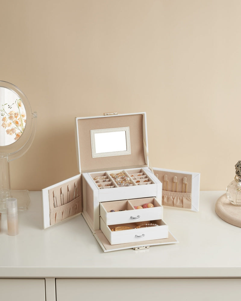 Open white jewellery box on a dressing table beside a round mirror and a perfume bottle, filled with rings and beads