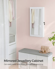 White mirrored jewellery cabinet mounted on a bedroom wall beside a padded bench and a handbag