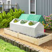 White greenhouse sitting on a wooden raised bed in a garden with flowers and a shed behind