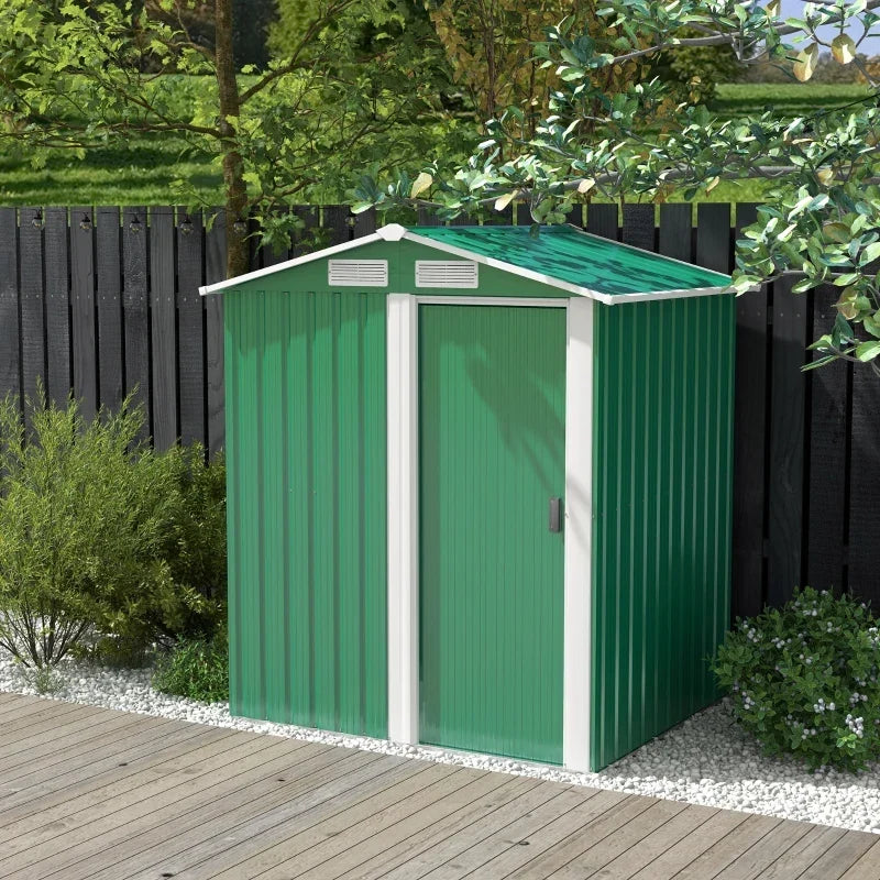 Green metal garden shed standing on decking in front of a dark painted fence and shrubs