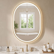 Gold oval LED mirror lit above a vanity with a window and bath reflected in it