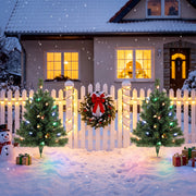Two lit trees beside a white picket fence outside a snowy house