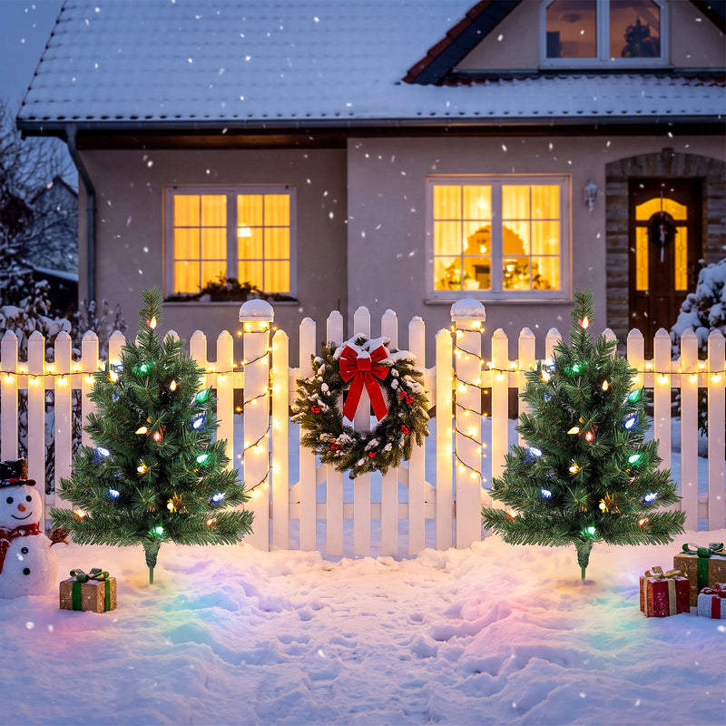 Two lit trees beside a white picket fence outside a snowy house