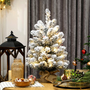 Lit slim flocked mini tree on a table beside a lantern and grey curtains