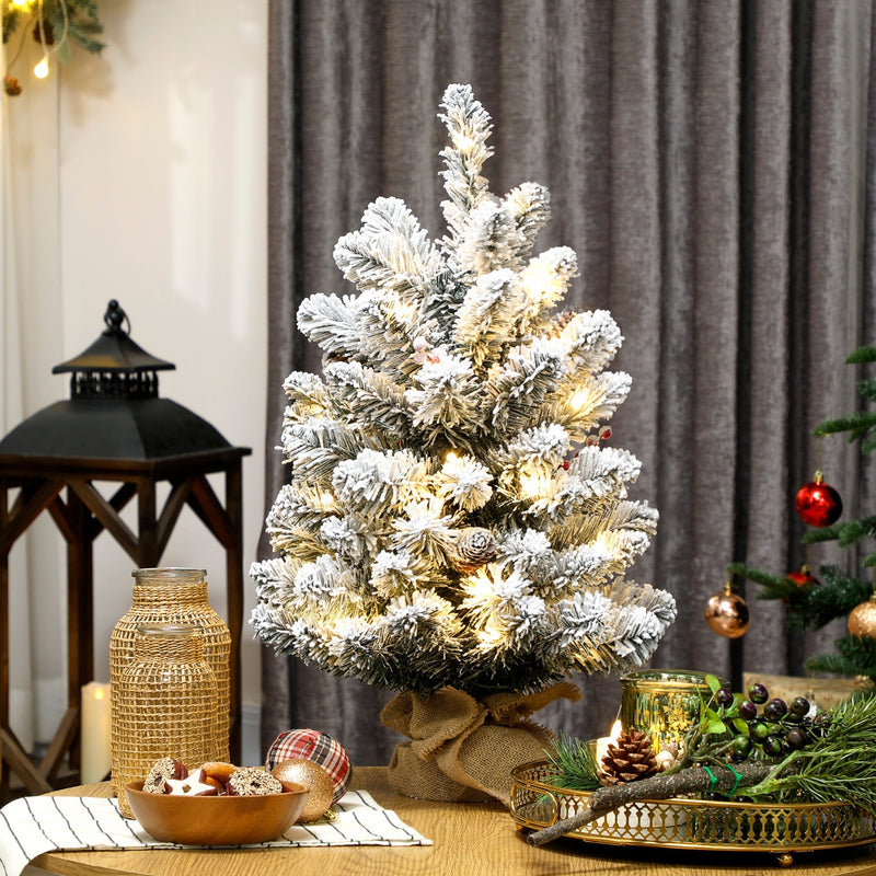 Lit slim flocked mini tree on a table beside a lantern and grey curtains