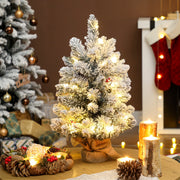 Lit slim flocked mini tree on a table beside red stockings, candles and a fireplace
