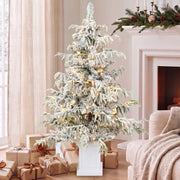 4ft snow flocked Christmas tree beside a fireplace with wrapped gifts around the pot