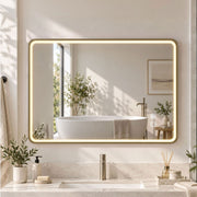 Gold-framed LED bathroom mirror lit above a vanity with a freestanding bath reflected in it