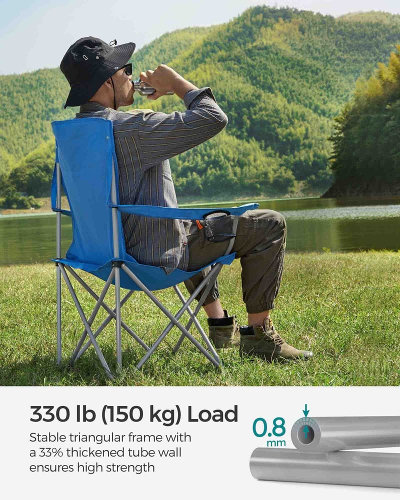 Man in a sun hat drinking while seated in the blue camping chair beside a lake