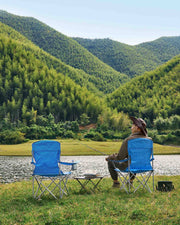 Two blue camping chairs set up on a lakeside lawn with a person fishing between them