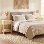 Oak bedside table next to a bed with a beige quilt and rug