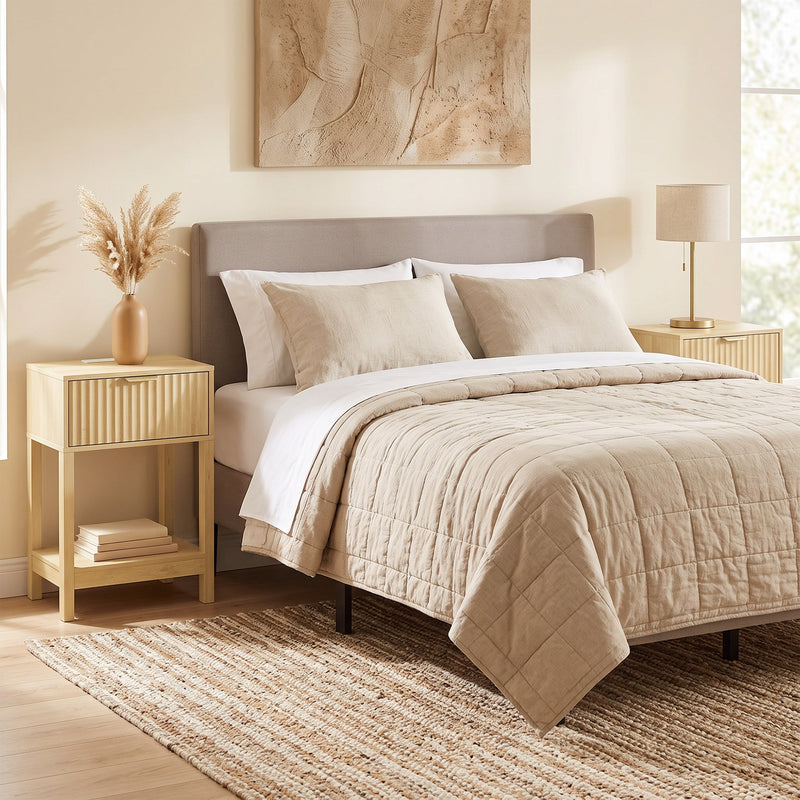 Oak bedside table next to a bed with a beige quilt and rug