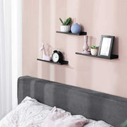 Black wall shelves on a pink bedroom wall above a bed, holding plants and a photo frame