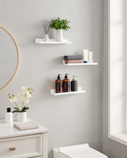 Three white ledge shelves in a bathroom holding toiletries, towels and flowers