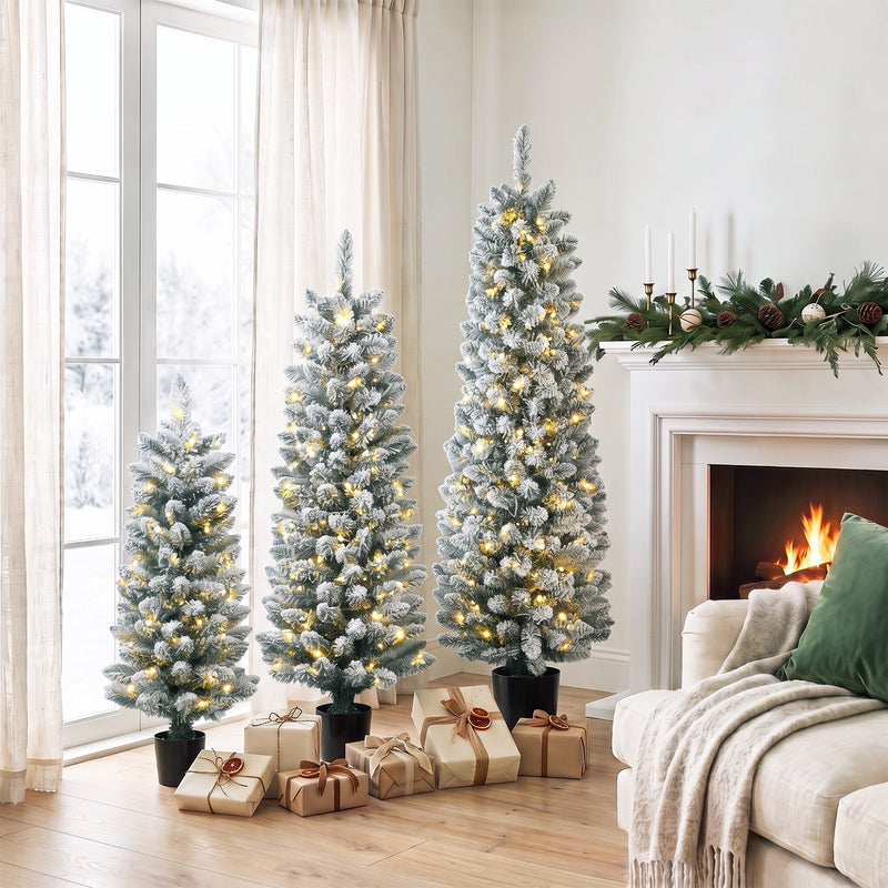 Three flocked Christmas trees with warm white lights by a window and fireplace with wrapped gifts