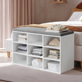 Lifestyle photo of the white shoe storage bench with grey cushioned seat placed at the foot of a bed, storing folded clothes and books