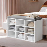 Lifestyle photo of the white shoe storage bench with grey cushioned seat placed at the foot of a bed, storing folded clothes and books