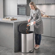 Woman scraping a plate into the open silver pedal bin in a grey kitchen