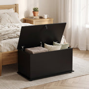 Black storage bench beside a wooden bed with a folded blanket and cushions in the open compartment