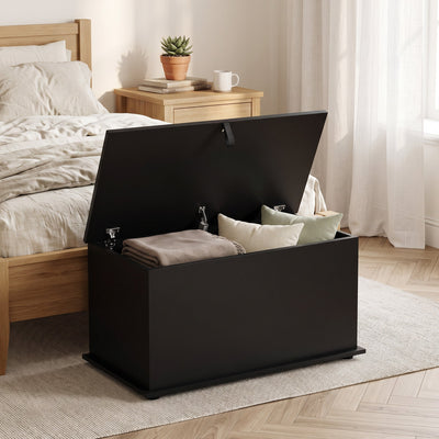 Black storage bench beside a wooden bed with a folded blanket and cushions in the open compartment