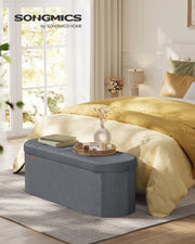 Slate grey ottoman bench used as an end-of-bed table with a tray, books and a vase in a bedroom.