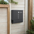 Ink black wall-mounted mailbox fixed to a pale timber fence beside trailing greenery
