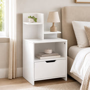 White bedside table beside a bed with a table lamp, plant and books