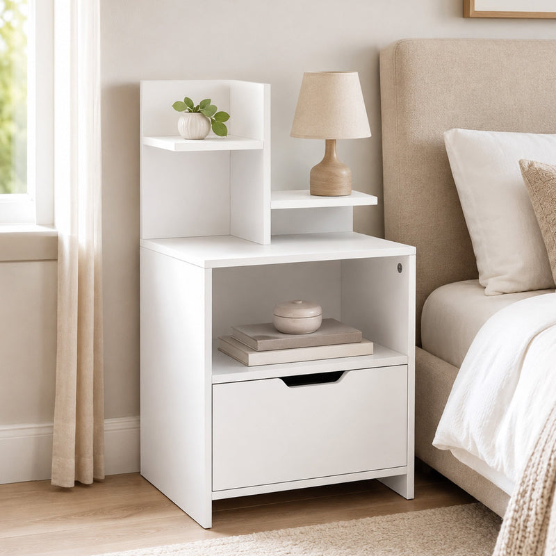 White bedside table beside a bed with a table lamp, plant and books