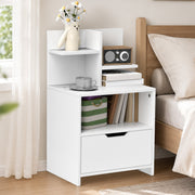 White bedside table beside a wooden bed with flowers, a radio and a cup on top