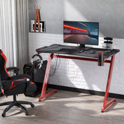 Red-framed gaming desk against a white brick wall with a monitor and gaming chair