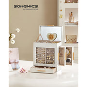 The Dressing Table Jewellery Organiser in white is open, showcasing a collection of earrings, necklaces, and rings. It includes a mirror inside the lid, a heart-shaped accessory, and elegant golden handles. Placed perfectly on the dressing table, it offers abundant storage against a backdrop of flowers and decor items.