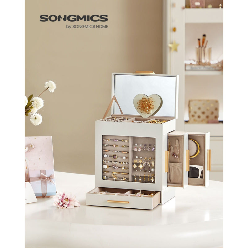 The Dressing Table Jewellery Organiser in white is open, showcasing a collection of earrings, necklaces, and rings. It includes a mirror inside the lid, a heart-shaped accessory, and elegant golden handles. Placed perfectly on the dressing table, it offers abundant storage against a backdrop of flowers and decor items.